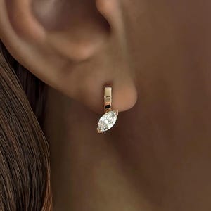 Può includere: Un orecchino color oro con un diamante taglio marquise. L'orecchino è un piccolo cerchio con un singolo pendente di diamante allungato. Il diamante è trasparente e riflette la luce. L'orecchino è indossato sull'orecchio.