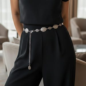 May include: A silver-toned belt with oval and circular decorative elements. The belt is worn over a black jumpsuit. The belt has a chain that hangs down from the center. The background shows a room with chairs and curtains.