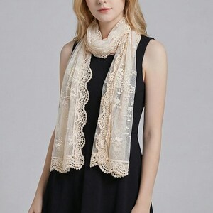 May include: A cream lace scarf with a floral design and scalloped borders. The scarf is draped around the neck and shoulders. The scarf is made of a sheer, lightweight material. The scarf adds a touch of elegance.
