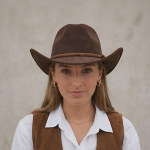 May include: A brown cowboy hat with a braided band. The hat is worn with a brown suede vest and a white collared shirt. The hat's brim is curved, and the overall style is Western.