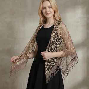 May include: A sheer, brown lace shawl with floral embroidery and a fringed edge. The shawl is draped over a black dress, creating a layered effect. The delicate design and warm color palette suggest an elegant accessory.