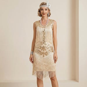 Peut inclure: Une robe de style flapper crème avec des détails de paillettes et de franges dorées. La robe sans manches présente un motif géométrique sur le corsage et une jupe à franges superposées. La modèle porte un bandeau et un collier de perles, évoquant une esthétique vintage.