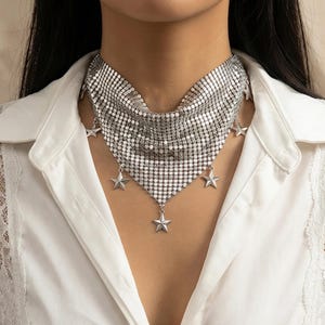 May include: A silver-toned choker necklace with a mesh design, featuring dangling star-shaped charms. The necklace is worn over a white collared shirt, creating a contrast between the shiny metal and the fabric.