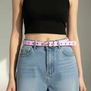 May include: A light pink belt with star-shaped cutouts and a silver star-shaped buckle. The belt is worn with blue jeans and a black top. The belt is approximately 1.25 inches wide.