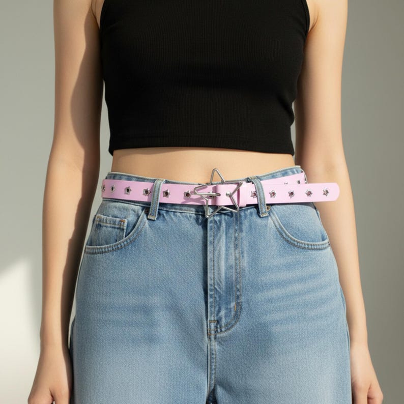 May include: A light pink belt with star-shaped cutouts and a silver star-shaped buckle. The belt is worn with blue jeans and a black top. The belt is approximately 1.25 inches wide.