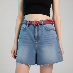 May include: A red leather belt with a silver star-shaped buckle and silver grommets. The belt is worn with denim shorts. The shorts are light blue and have a frayed hem.