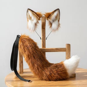May include: A set of faux fur fox ears on a thin headband and a matching long fox tail with a black adjustable strap. The ears are brown with white inner fur and dark tips. The tail is brown with a white tip, featuring long, soft synthetic fibres.