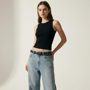 May include: A black tank top with a round neckline. The top is paired with light blue jeans and a black belt with a star-shaped buckle. The belt also has silver-colored grommets. The model is standing in front of a white background.
