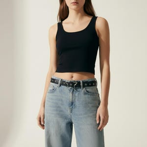 May include: A black crop top with a scoop neck is paired with light blue denim jeans. A black belt with a silver star buckle and silver stud accents completes the outfit. The overall style is casual and modern.