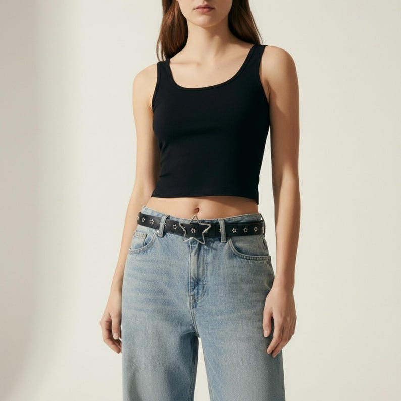 May include: A black crop top with a scoop neck is paired with light blue denim jeans. A black belt with a silver star buckle and silver stud accents completes the outfit. The overall style is casual and modern.