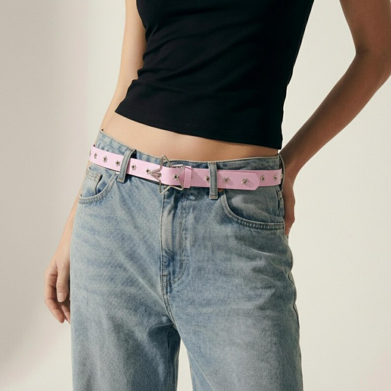 May include: A light pink belt with silver-tone star-shaped studs and a silver buckle. The belt is worn with light blue denim jeans and a black top. The belt is approximately 1.25 inches wide.