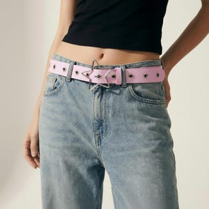 May include: A light pink belt with a star-shaped buckle and silver grommets. The belt is worn with light wash blue jeans. The belt is approximately 1.5 inches wide.