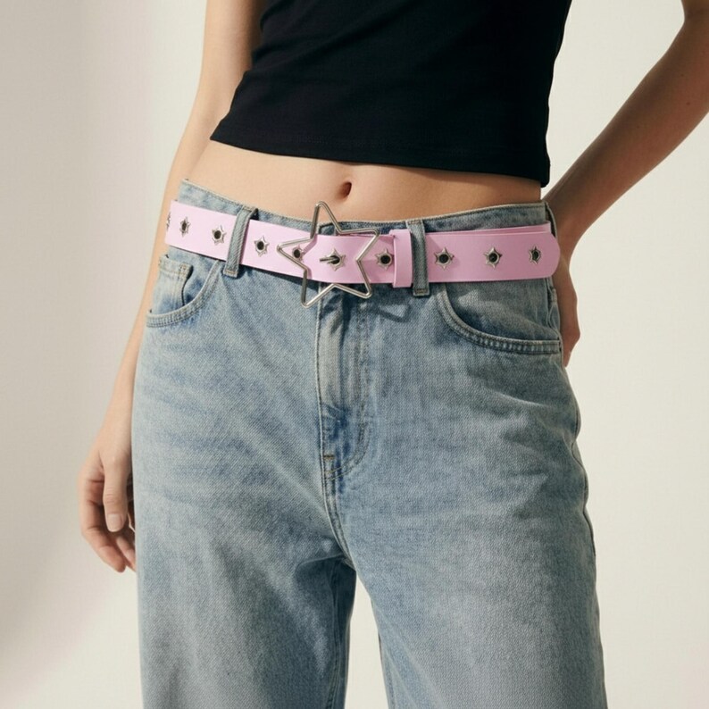 May include: A light pink belt with a star-shaped buckle and silver grommets. The belt is worn with light wash blue jeans. The belt is approximately 1.5 inches wide.