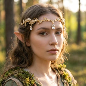 May include: A gold-toned metal headpiece featuring a delicate vine and leaf design adorned with small clear crystals. The accessory includes three dangling teardrop-shaped clear crystals, with one centred on the forehead and two on the sides.