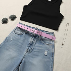 May include: A flat lay of a black crop top, light blue denim jeans with distressed details, a pink belt with star-shaped buckle, silver sunglasses, and a silver necklace with a pendant. The belt has small star-shaped studs.