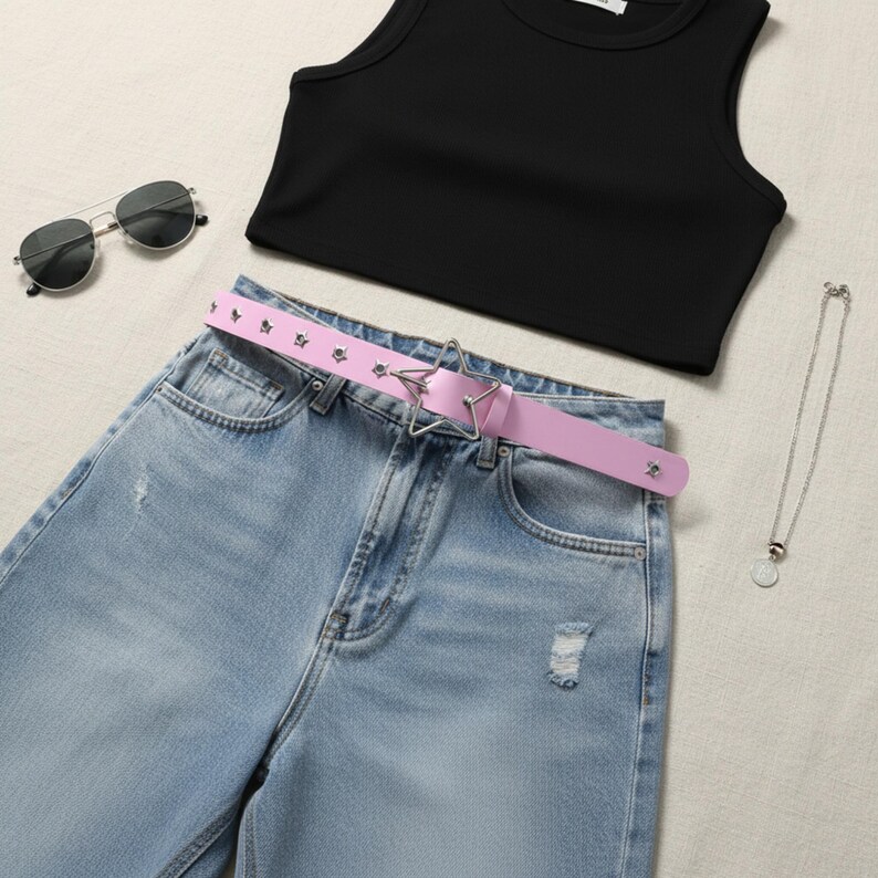 May include: A flat lay of a black crop top, light blue denim jeans with distressed details, a pink belt with star-shaped buckle, silver sunglasses, and a silver necklace with a pendant. The belt has small star-shaped studs.