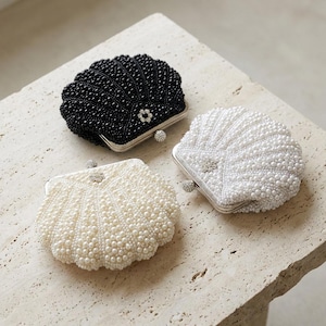 May include: Three shell-shaped beaded clutches in black, white, and cream. Each clutch has a silver clasp and a decorative embellishment. The clutches are displayed on a textured surface, highlighting their unique designs and textures.