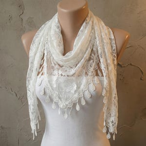 Puede incluir: Bufanda de encaje blanco sobre un maniquí. La bufanda presenta un delicado diseño floral y bordes en forma de lágrima. La tela es suave y transparente, ideal para añadir un toque de elegancia.