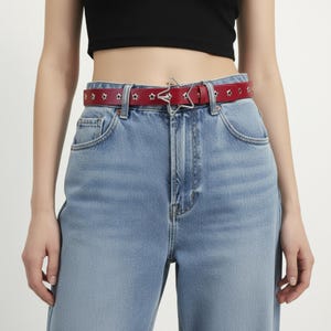 May include: A red leather belt with star-shaped cutouts and a silver star-shaped buckle. The belt is worn with light blue denim jeans. The belt is approximately 1.25 inches wide.