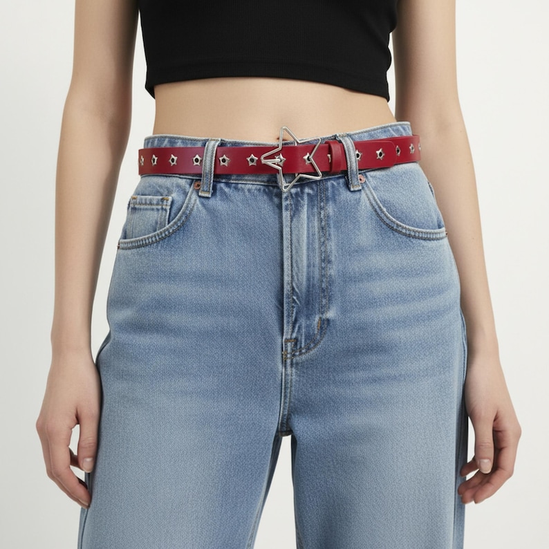 May include: A red leather belt with star-shaped cutouts and a silver star-shaped buckle. The belt is worn with light blue denim jeans. The belt is approximately 1.25 inches wide.