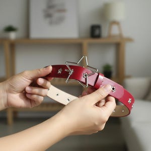 May include: A red leather belt with a silver star-shaped buckle. The belt has silver star-shaped studs and is held up by a person's hands. The belt is approximately 1.25 inches wide.