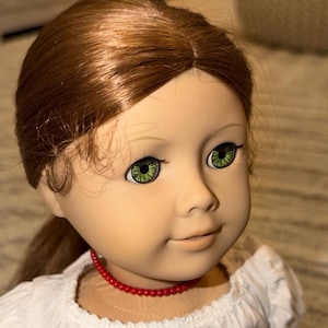 Puede incluir: Una muñeca con cabello castaño rojizo recogido en una cola de caballo, ojos verdes y un collar de cuentas rojas. La muñeca lleva una blusa blanca con un escote con volantes. La cara de la muñeca tiene una expresión suave.