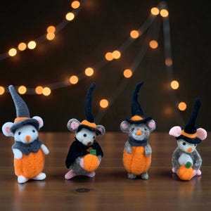 May include: Four Halloween-themed felt mice figurines. Each mouse wears a black witch hat with an orange band and a matching orange outfit. Some mice hold small orange pumpkins. The background features blurred string lights.