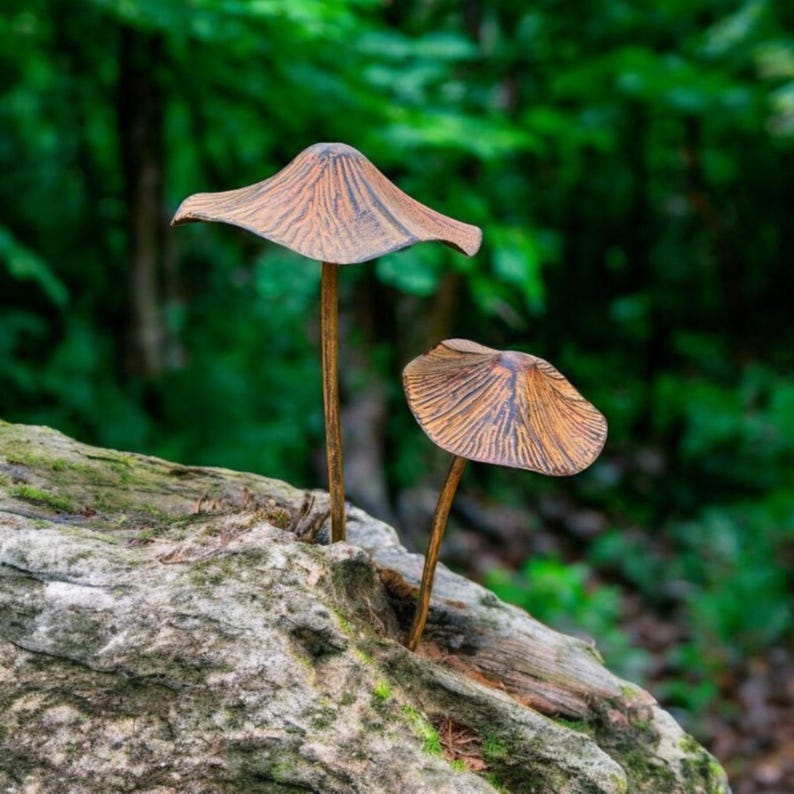 May include: Two bronze-colored mushroom sculptures with textured caps and slender stems emerge from a weathered, moss-covered log. The background features blurred green foliage, creating a natural, earthy scene.
