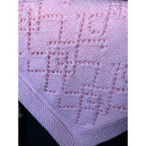 May include: A light pink knitted blanket featuring a textured geometric pattern created with raised stitches. The design consists of interconnected squares and lines formed by knit and purl stitches, creating a soft, uniform fabric surface.
