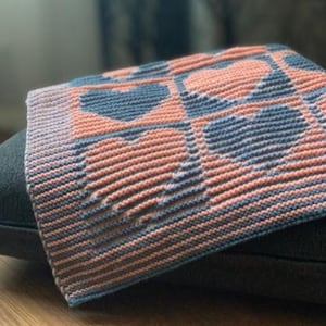 May include: A knitted blanket with a heart pattern in shades of coral and blue. The blanket is folded over a dark surface, showcasing the textured stitch work and color contrast. The blanket's design features repeating heart shapes.