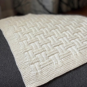May include: Cream-colored knitted blanket with a woven basket weave pattern. The blanket is made of soft yarn and has a textured surface. The blanket is folded over a dark gray surface.