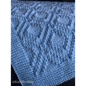 May include: A light blue knitted blanket featuring a textured diamond pattern created with raised stitches. The fabric has a consistent garter stitch border and a repeating geometric design. The text @KnitHitShop is visible in the corner.