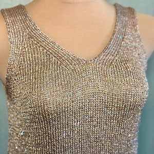 May include: A sleeveless, gold-colored knit top with a V-neck. The fabric has a shimmering, textured appearance with subtle iridescent accents. The top is designed with a close fit and a classic silhouette.