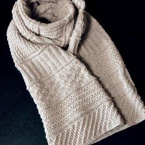 May include: A beige knitted scarf with a variety of textured patterns. The scarf is folded, displaying different stitch designs, including ribbed and diamond patterns. The scarf is made of a soft, warm material, ideal for colder weather.