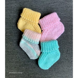 May include: Four hand-knitted baby booties featuring folded cuffs. One is solid yellow, one is solid pink, one is solid turquoise, and one is a variegated blend of light pink, light blue, and white. The text @KnitHitShop is visible in the corner.