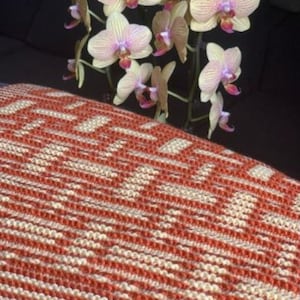 May include: A knitted blanket with a geometric pattern in red and cream colors. The blanket is folded and displayed with a bouquet of light pink and yellow orchids. The blanket is a cozy home decor item.