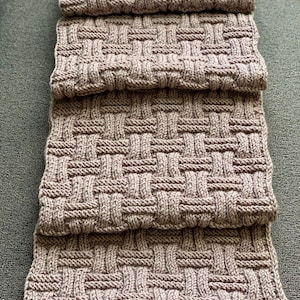 May include: A folded, woven-style scarf in a neutral taupe color. The scarf is made of a soft, knitted material with a basketweave pattern. The scarf is folded in thirds, showing the texture and design. The background is a solid, muted green.