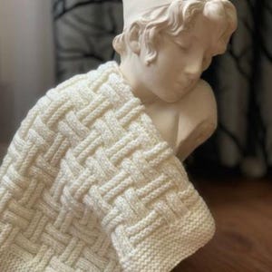 Puede incluir: Manta de punto color crema con un patrón de tejido de cesta, drapeada sobre un busto clásico. La textura de la manta es visible, con un borde acanalado. El busto es de color beige claro. La manta es un elemento de decoración acogedor.