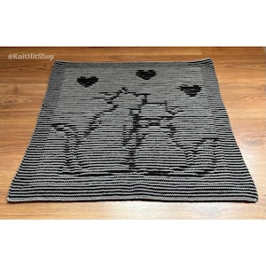 Puede incluir: Una manta cuadrada de ganchillo con un patrón de rayas horizontales negras y grises. El diseño muestra la silueta central de dos gatos frente a frente con tres corazones pequeños encima. El texto KnitHitShop es visible en la esquina.