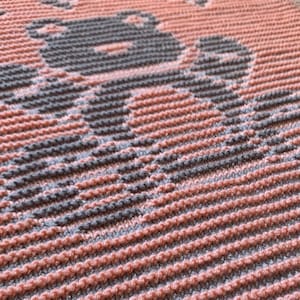 May include: Close-up of a knitted blanket with a pink and gray pattern. The pattern is a repeating design of horizontal stripes.
