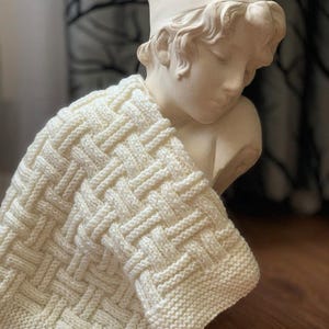 以下が含まれることがあります： クリーム色のバスケット織りの模様のニットブランケットが、古典的な胸像に掛けられています。 ブランケットの質感は、詳細な織り方で確認できます。 胸像は薄いベージュ色で、背景はぼやけています。