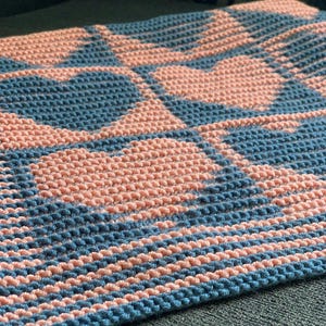May include: A handmade crochet blanket featuring a repeating heart pattern. The blanket is made with a textured stitch in shades of light pink and blue. The blanket is folded, showing the pattern clearly.