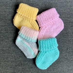 Può includere: Quattro paia di scarpine per neonati lavorate a maglia in colori pastello: giallo, rosa, blu e un paio multicolore con strisce rosa, blu e grigie. Le scarpine hanno polsini ripiegati e sono disposte su una superficie grigia strutturata.