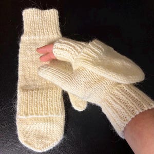 Könnte beinhalten: Cremefarbene Strickfäustlinge und ein passender Ärmel. Die Fäustlinge haben ein fingerloses Design mit einer Klappe, die die Finger bedecken kann. Der Ärmel ist lang und oben und unten gerippt. Die Fäustlinge und der Ärmel sind aus weichem Garn.