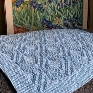 Puede incluir: Una manta de punto azul claro con un patrón geométrico y un borde liso. La manta se muestra sobre una superficie oscura, con una pintura enmarcada de flores azules en el fondo. El nombre de la tienda de Etsy @KnitHitShop es visible.