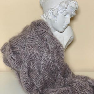 May include: A soft, gray knitted scarf draped over a white bust of a classical figure. The scarf has a textured, cable-knit design. The bust is of a person with closed eyes and a serene expression. The background is a light beige color.