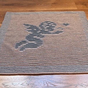 May include: A square, hand-crocheted rug featuring a detailed design of a cherub holding a heart. The cherub is in a dark gray color, set against a tan and gray striped background. The rug has a gray border.