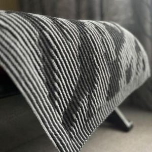 Puede incluir: Una manta de punto a rayas blancas y negras sobre un banco. La manta tiene un patrón texturizado y acanalado. La imagen está tomada en interiores con luz natural. El nombre de la tienda, @KnitHitShop, es visible en la esquina inferior izquierda.