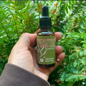 Op de afbeelding: Een glazen fles met een druppelaar gevuld met een heldere vloeistof. De fles heeft een groen etiket met de tekst "Supernatural Radiant Glow Pure Natural Beauty Elixir".