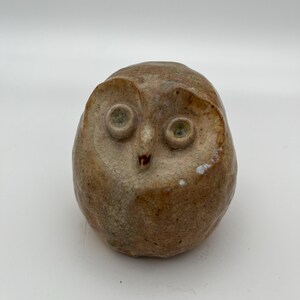 May include: A small ceramic owl figurine with a brown and white glaze. The owl has large, round eyes and a small beak.
